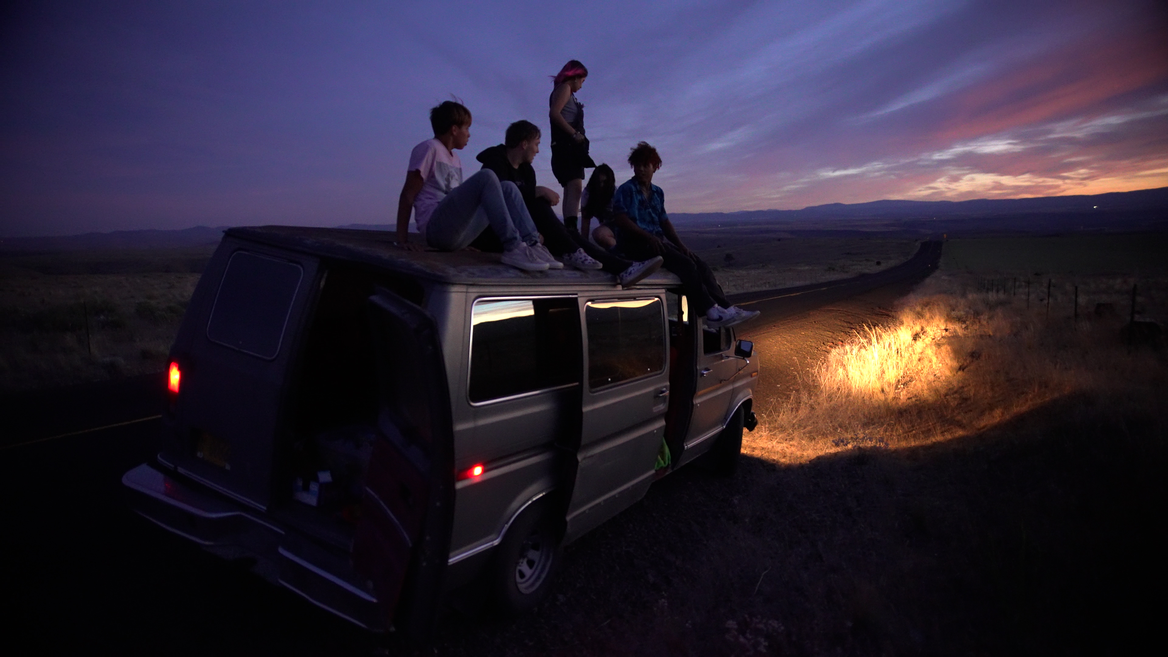 A group of teenagers sitting on the roof of a van at night time. It is facing away from the camera with its headlights pointing towards the horizon.
